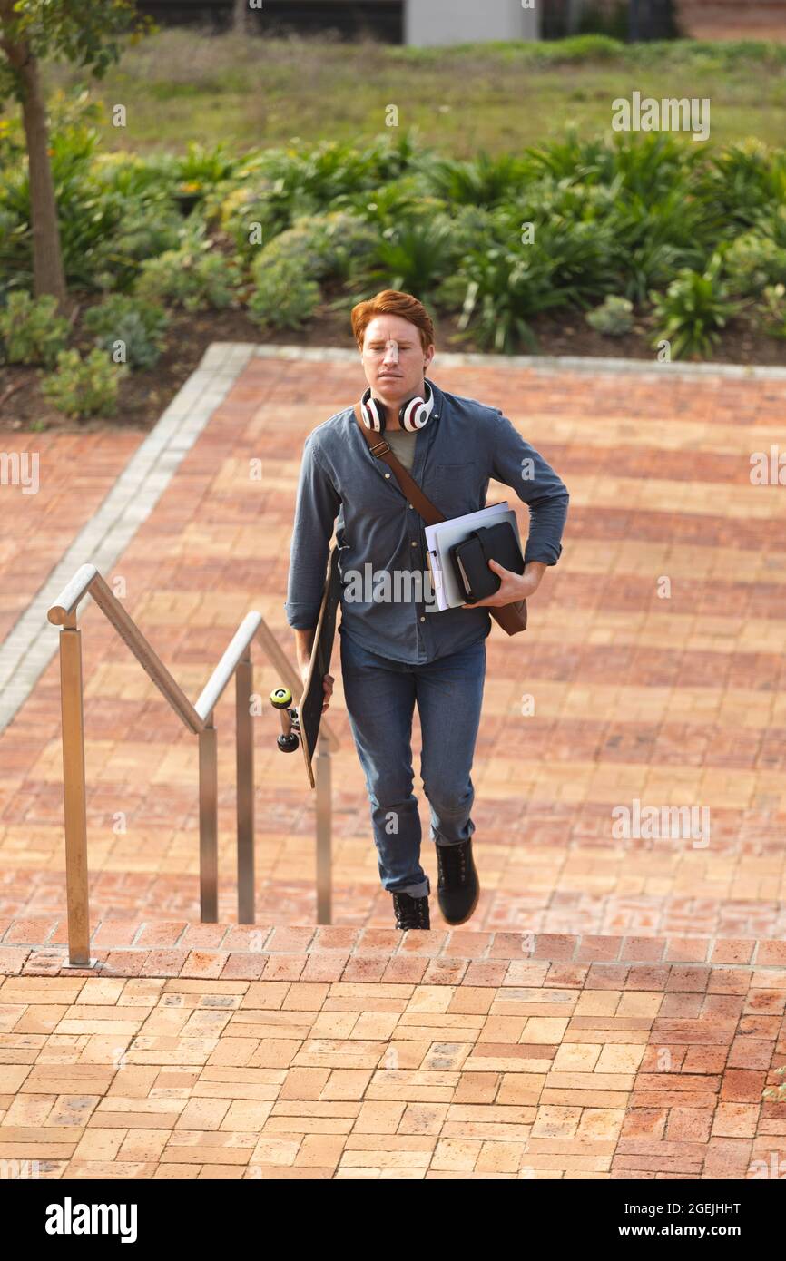 Serious caucasian male business creative walking upstairs holding notebooks and skateboard Stock Photo