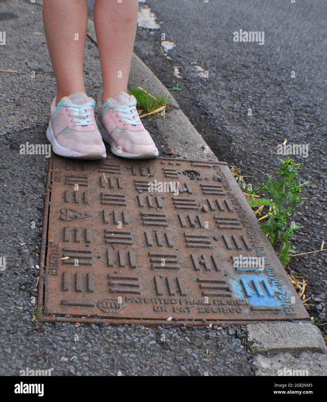 Man hole cover on the edge of a pavement. A blue dot of spray paint on the corner indicates it has been inspected. A child is standing at the edge Stock Photo