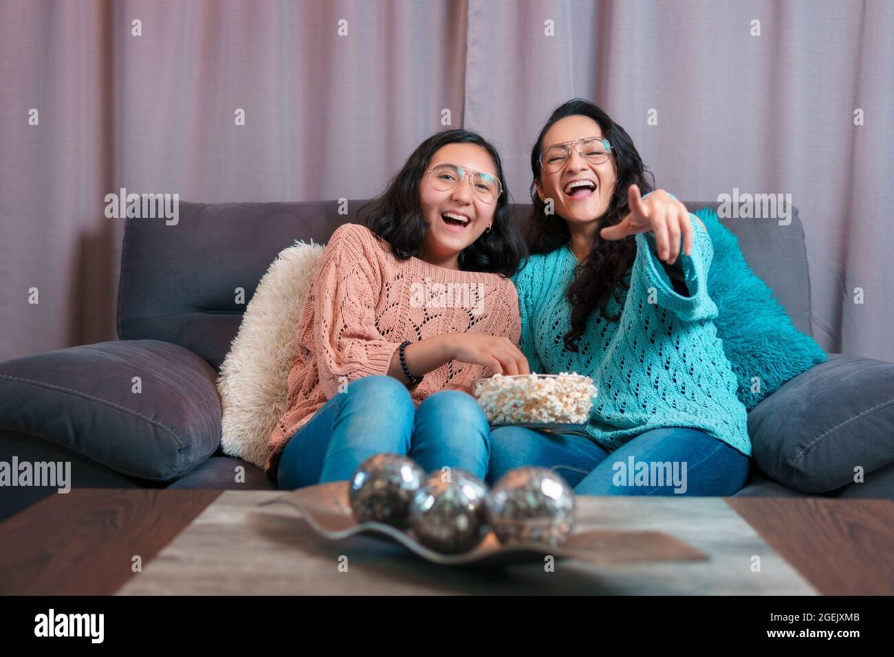 Horizontal view of a Mom and daughter watching a movie at home, they eat popcorn, they both laugh very funny and make funny expressions Stock Photo
