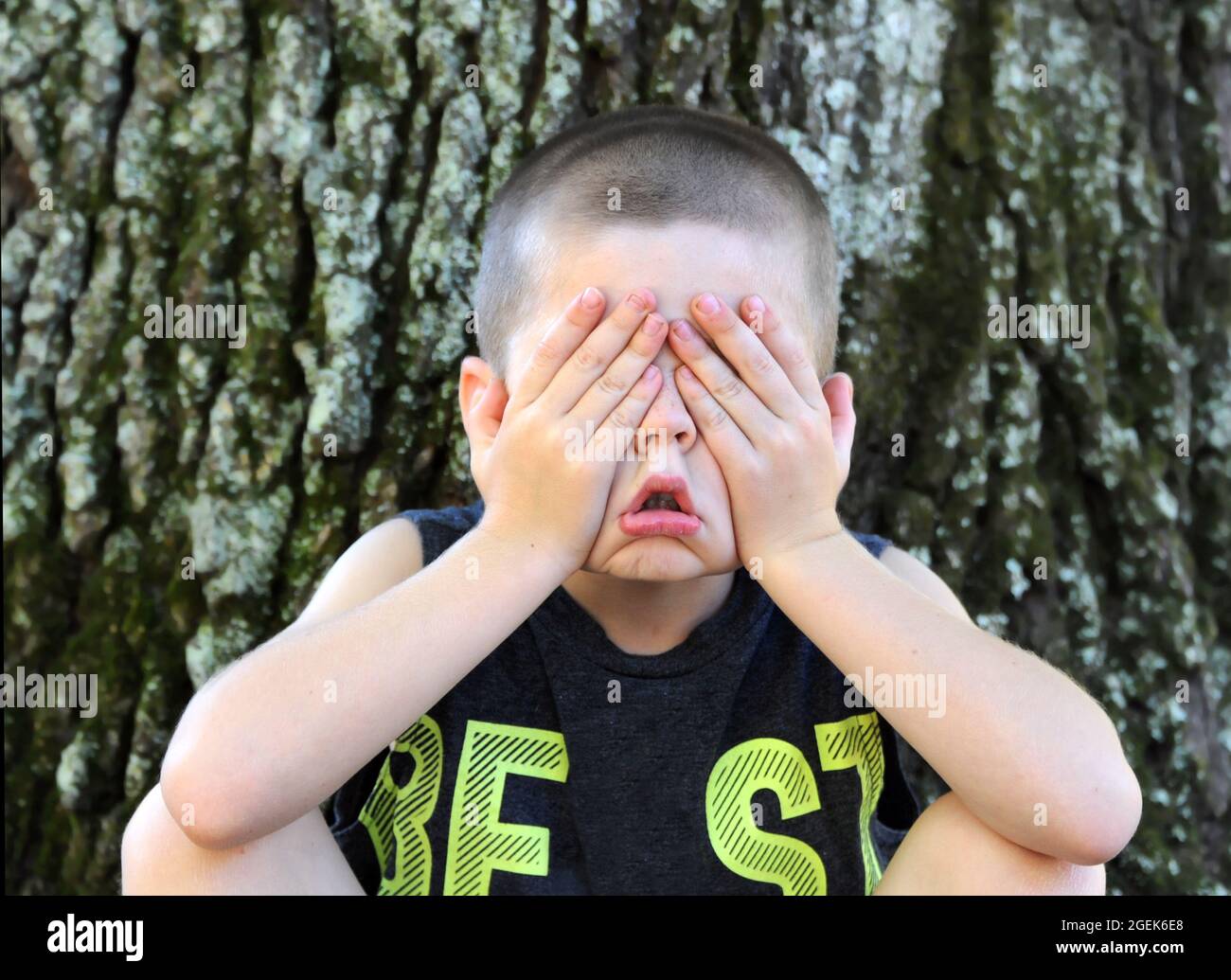Little boy sitting outdoors against a tree, hids his eyes so he can't