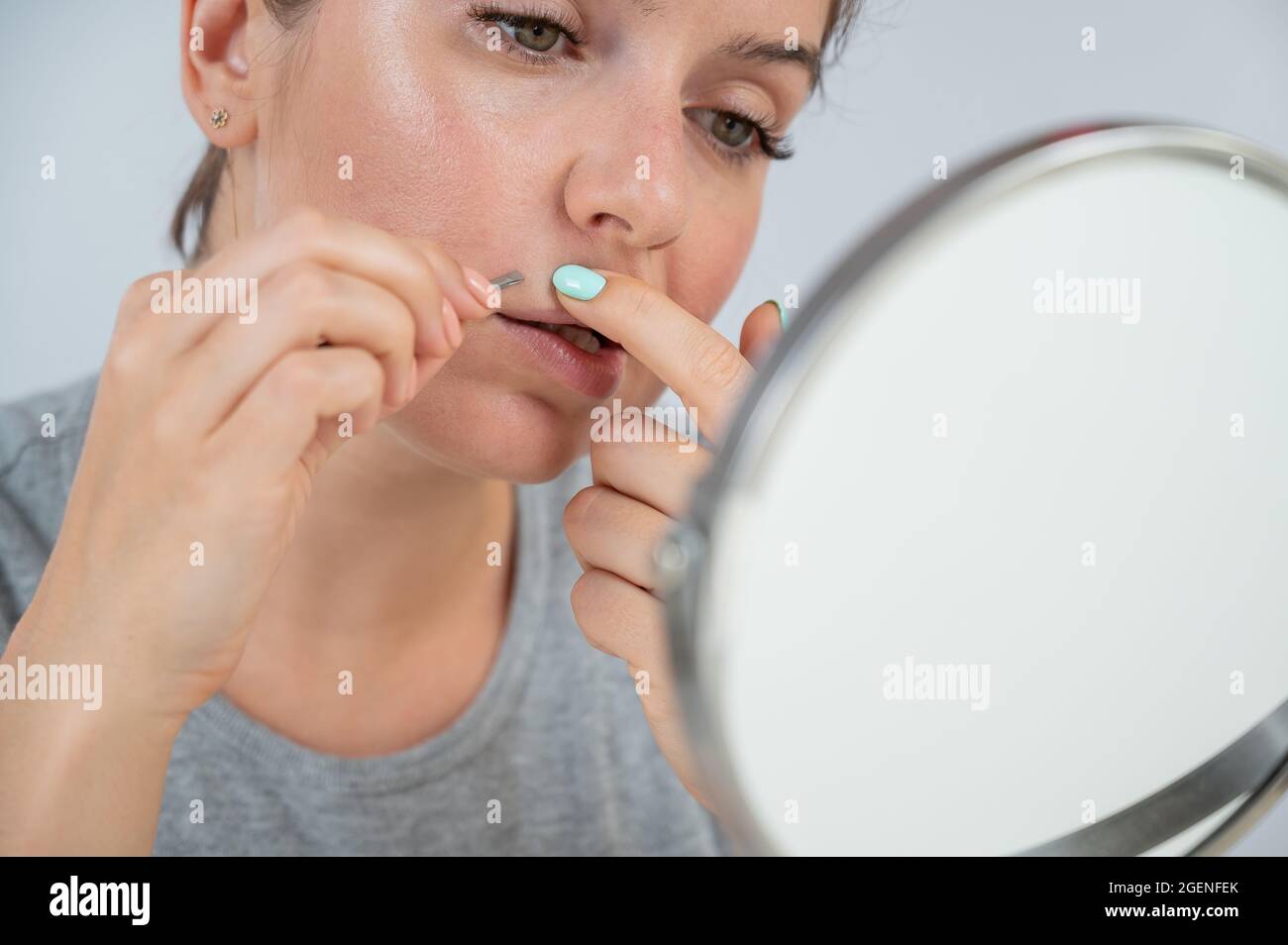 Caucasian woman looks in the mirror and removes the mustache herself with the help of tweezers. Hirsutism. Stock Photo