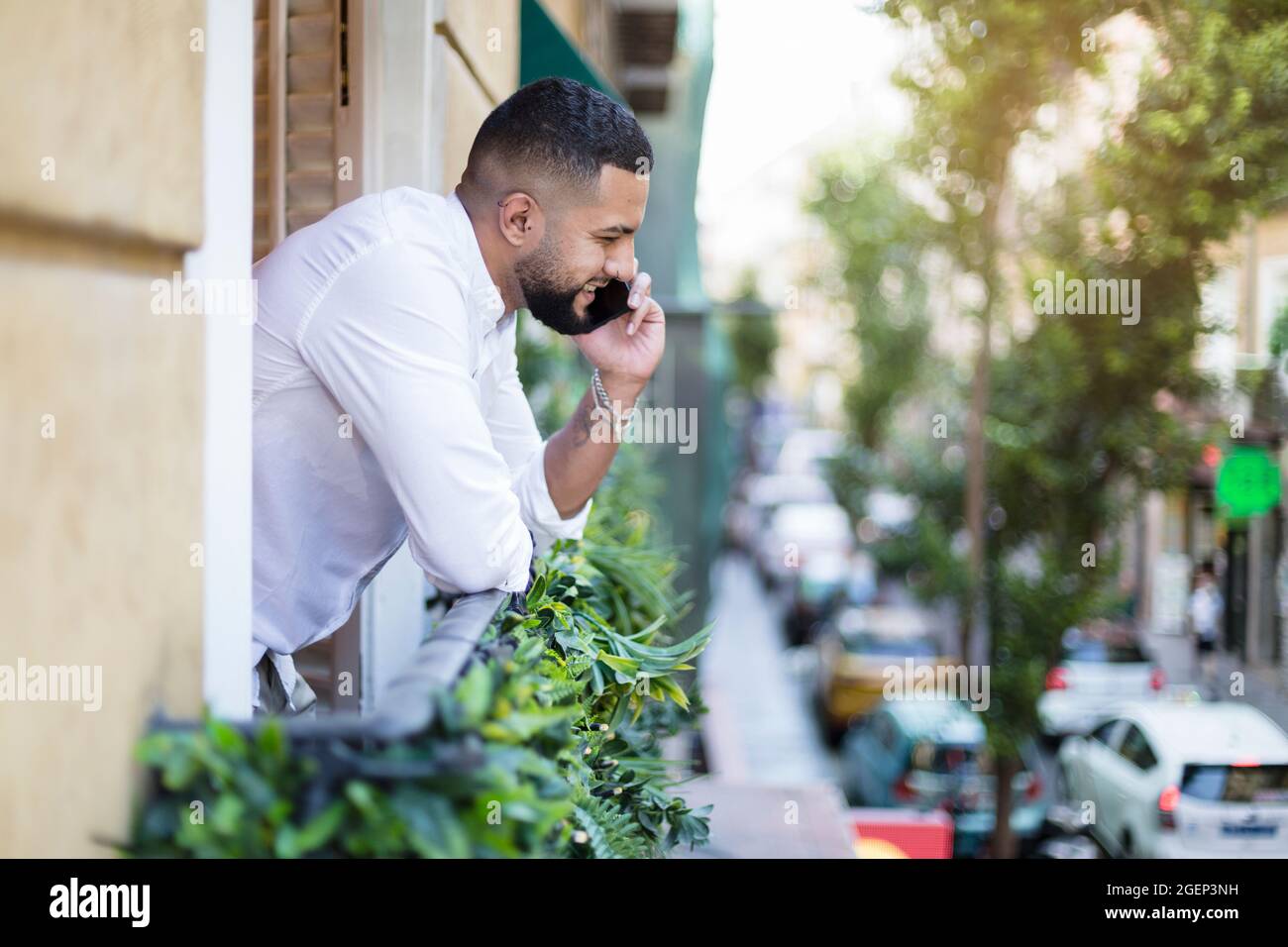 Latin guy talking on mobile phone leaning out of apartment balcony. Space for text. Stock Photo