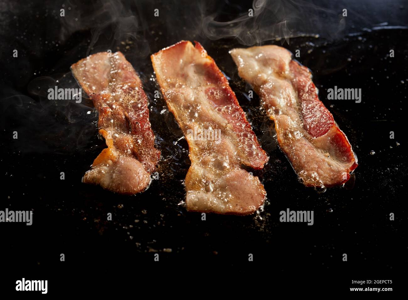 Closeup shot of delectable bacon strips cooking in oil on a hot black surface Stock Photo