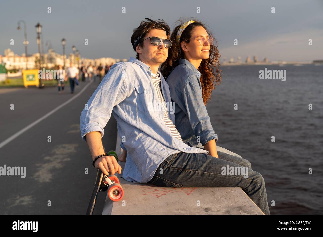 Stylish couple sit on pier at seaside enjoy sunset. Happy man and woman looking at river or sea Stock Photo