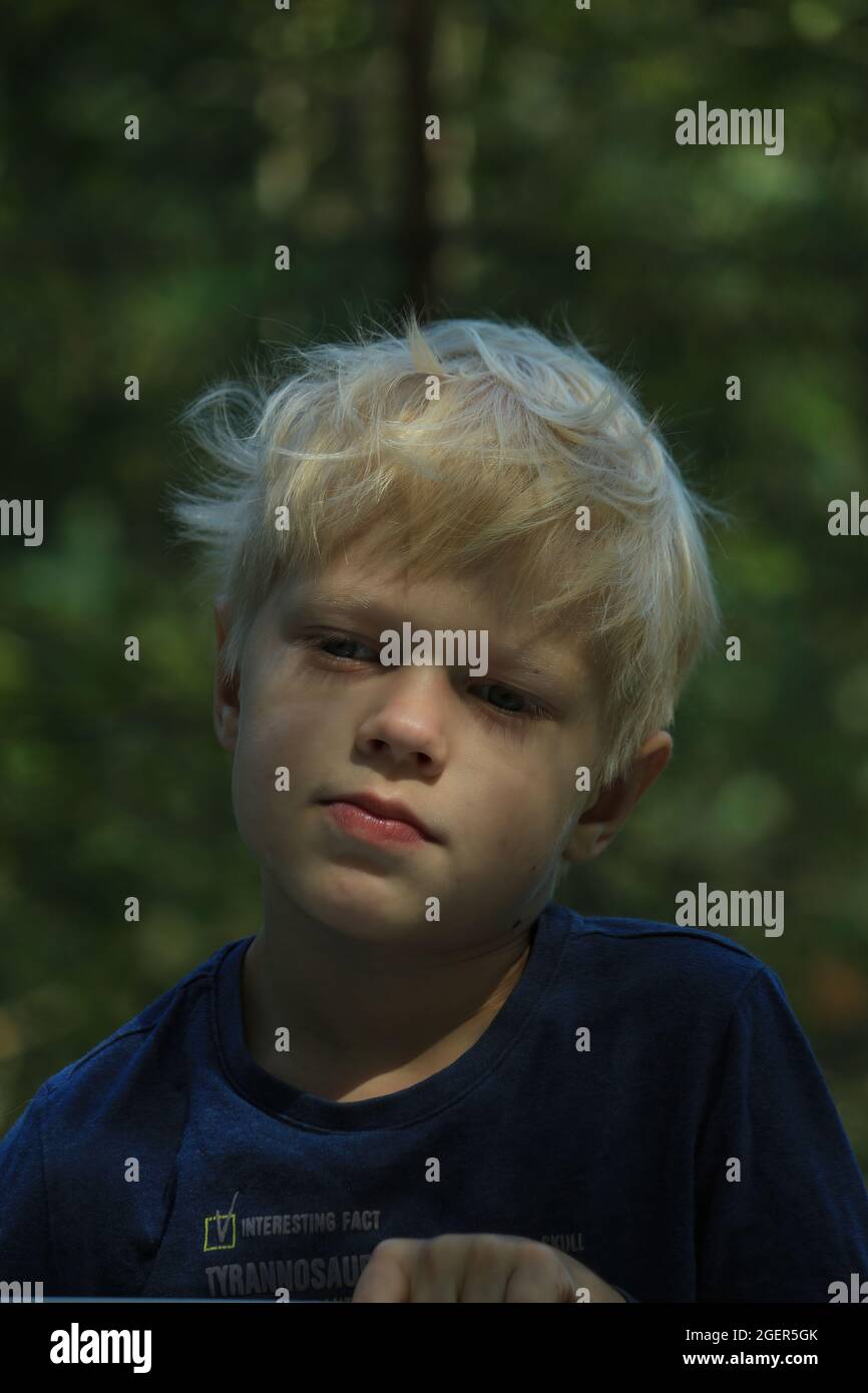Portrait of a blond boy with tousled hair outdoors on the background of a summer green forest. Stock Photo