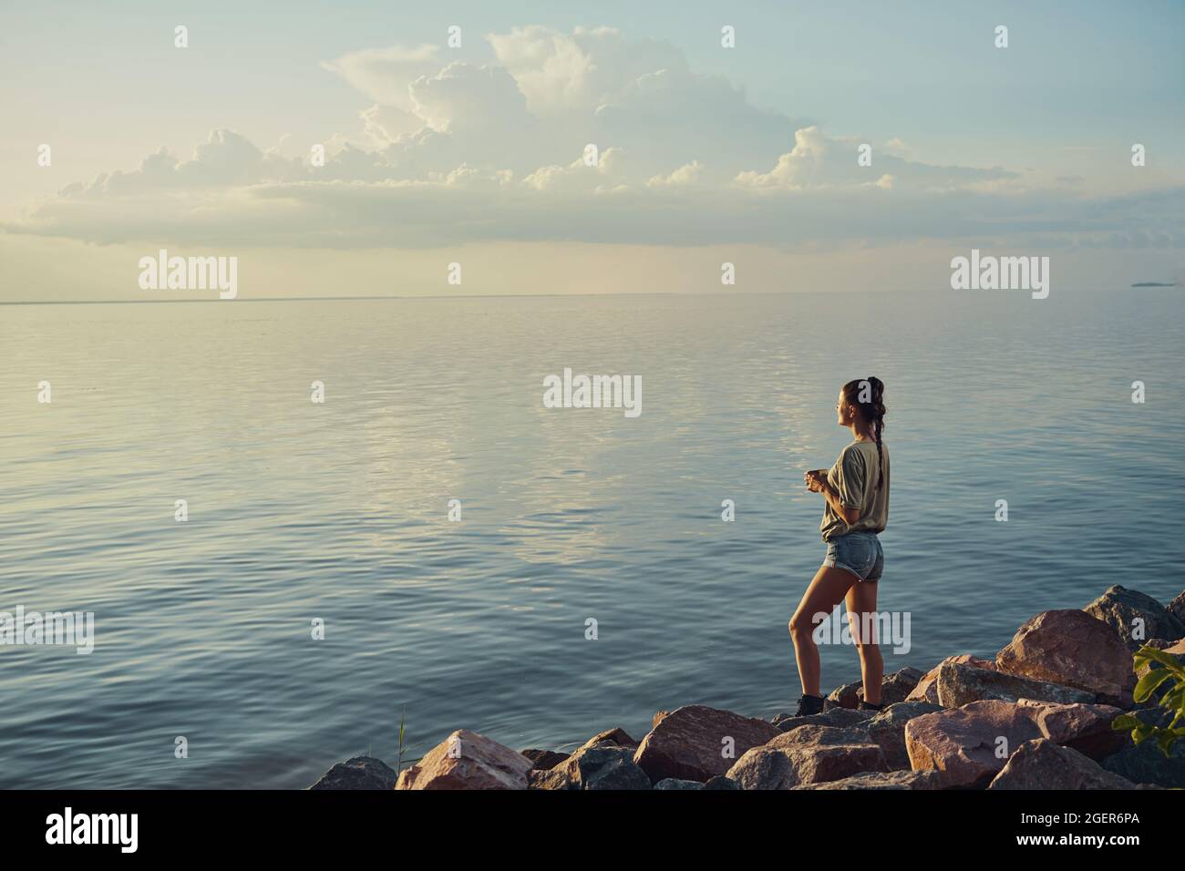 Adult standing on rocks with teacup and watching setting sun Stock Photo