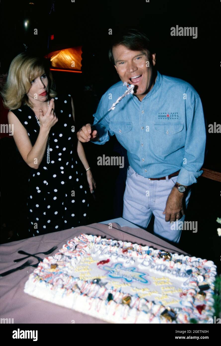 Glen Campbell pretends to lick the knife after cutting into his birthday cake at his surprise 60th birthday celebration on April 21, 1996 in Branson, Missouri. His wife, Kim looks on after sampling the icing on her fingers. Glen Campbell's actual birth date was April 22, 1936 Stock Photo