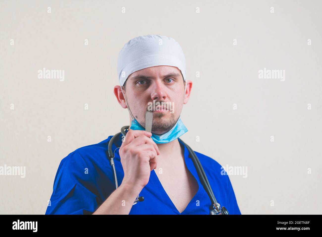 Young doctor portrait in blue suit, stethoscope and protective breathing mask with spatula in hand near his mouth close up isolated Stock Photo