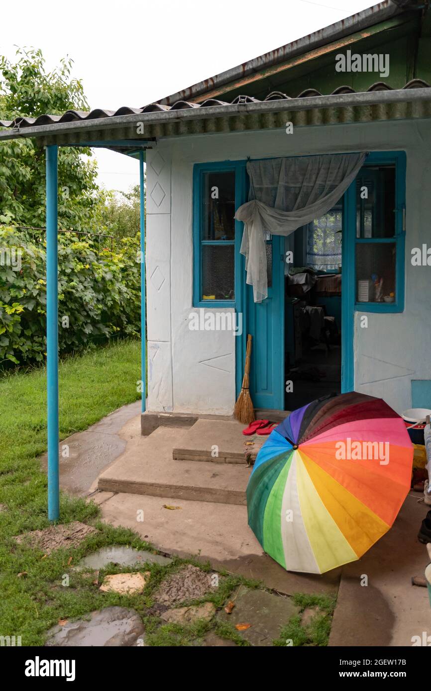 Wet umbrella of striped rainbow on grunge porch of farmhouse with opened door. Rustic scene in rain weather Stock Photo