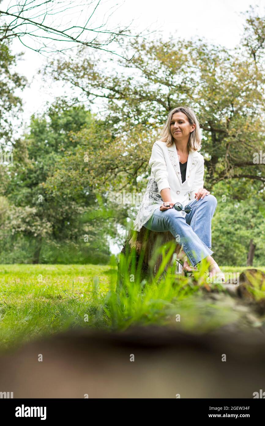 Woman posing sitting on a wooden log in a park in Asturias, Spain.The girl is wearing jeans, a white jacket and transparent shoes.Photo with a blur in Stock Photo
