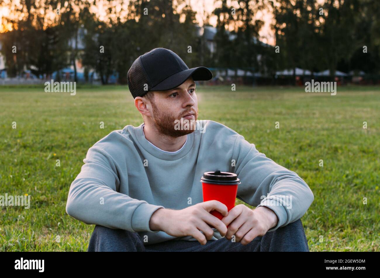Young man drinking coffee from a plastic cup on the grass thoughtfully Stock Photo