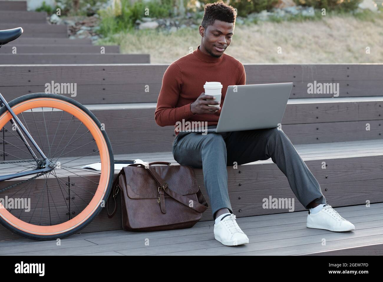 Young African student or freelancer with drink looking at laptop screen while sitting on staircase Stock Photo