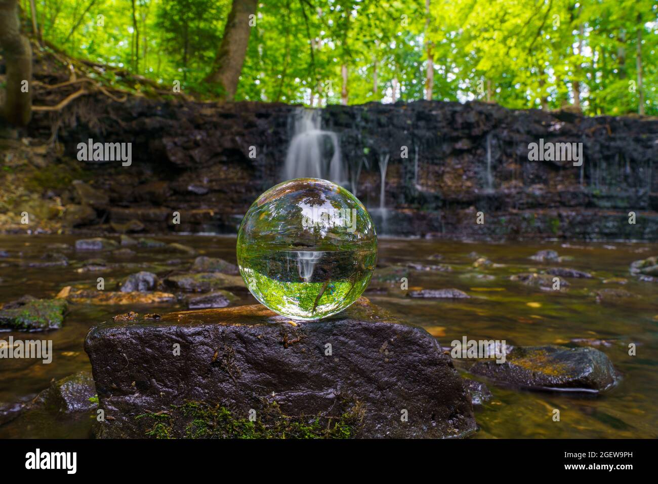 The glass ball stands on a square stone and through the glass ball you can see a waterfall in the air with your feet in the background, a fuzzy waterf Stock Photo