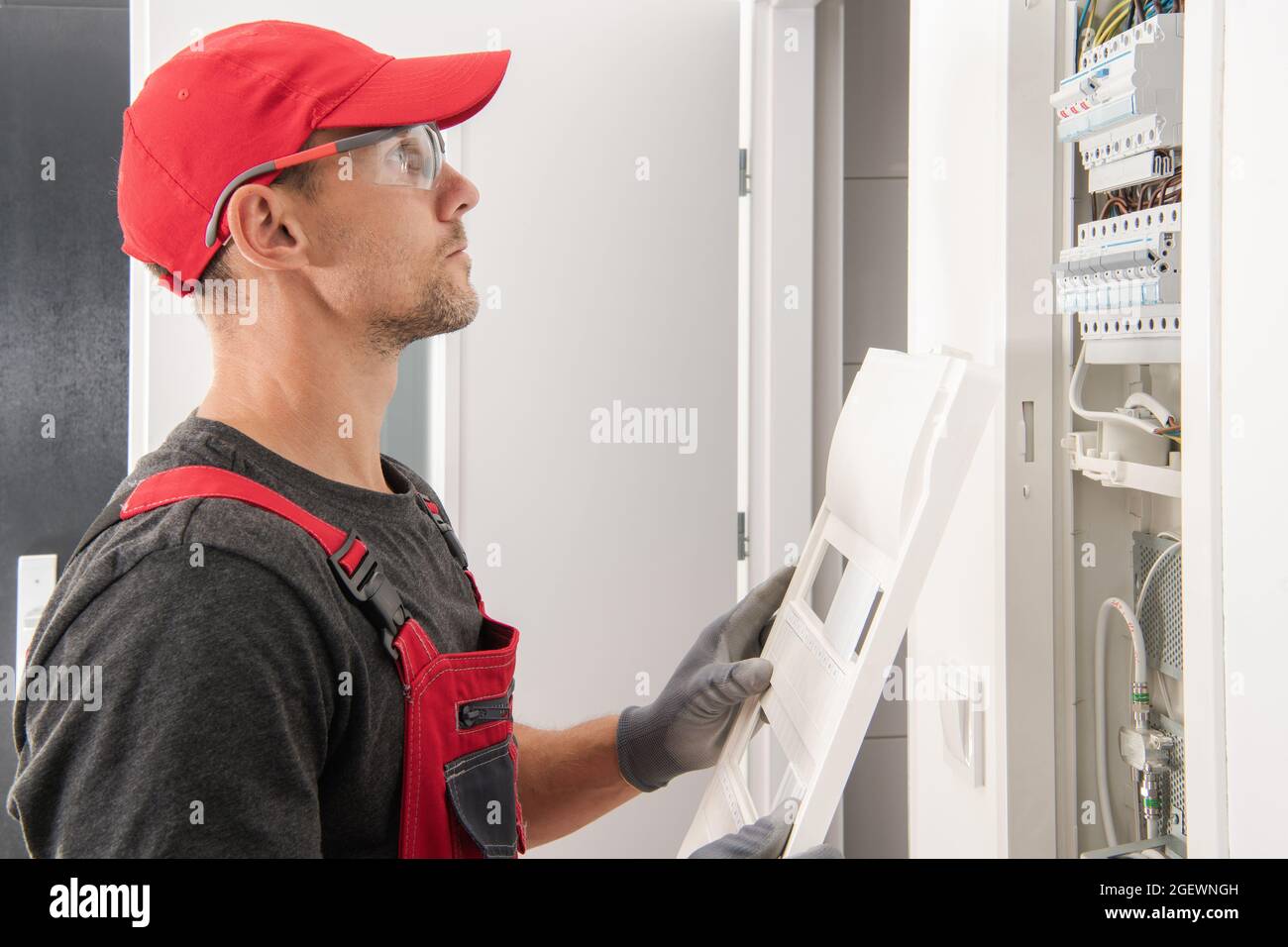 Residential Electric Systems. Professional Caucasian Electrician in His 40s Wearing Red Uniform and Safety Glasses Checking Residential Apartment Fuse Stock Photo