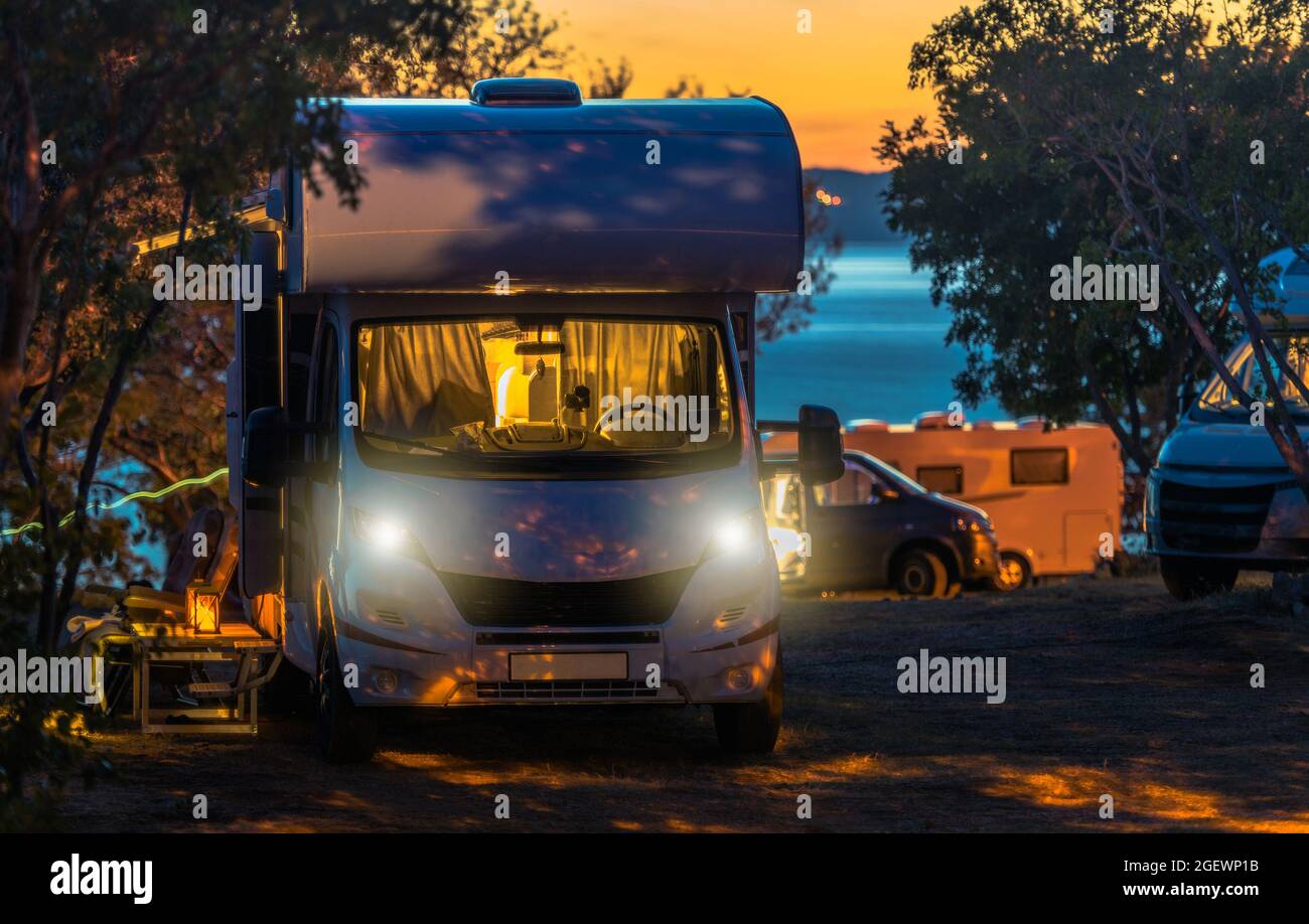Scenic RV Park Campsite Sunset. Modern Camper Vans Prepared For Overnight Stay. Last Call of Light Over the Sea in Background. Stock Photo