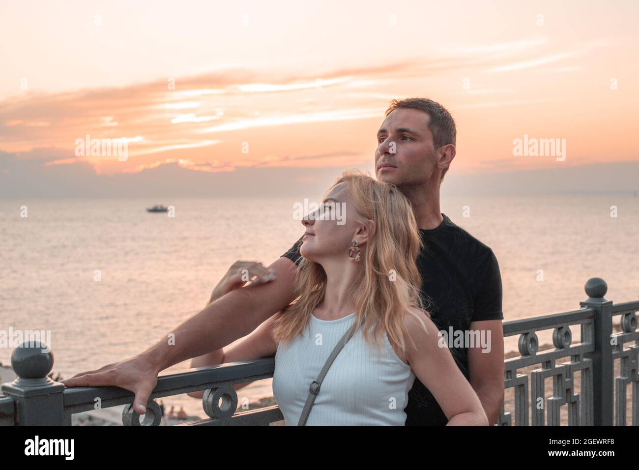 A man and a woman stand embracing on the embankment and look at the sea at sunset. Millennial generation. Stock Photo