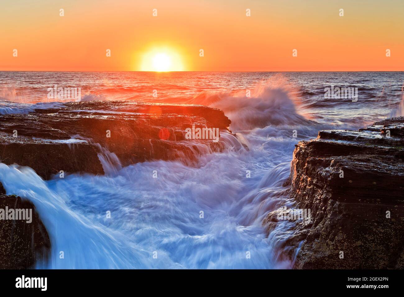 Wet sandstone rocks lit by Rising sun and Pacific ocean waves in scenic seascape - Sydney Northern beaches. Stock Photo