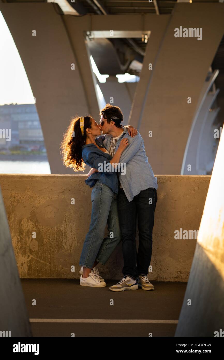 Romantic couple of young male and female embracing and kissing at sunset standing under urban bridge Stock Photo