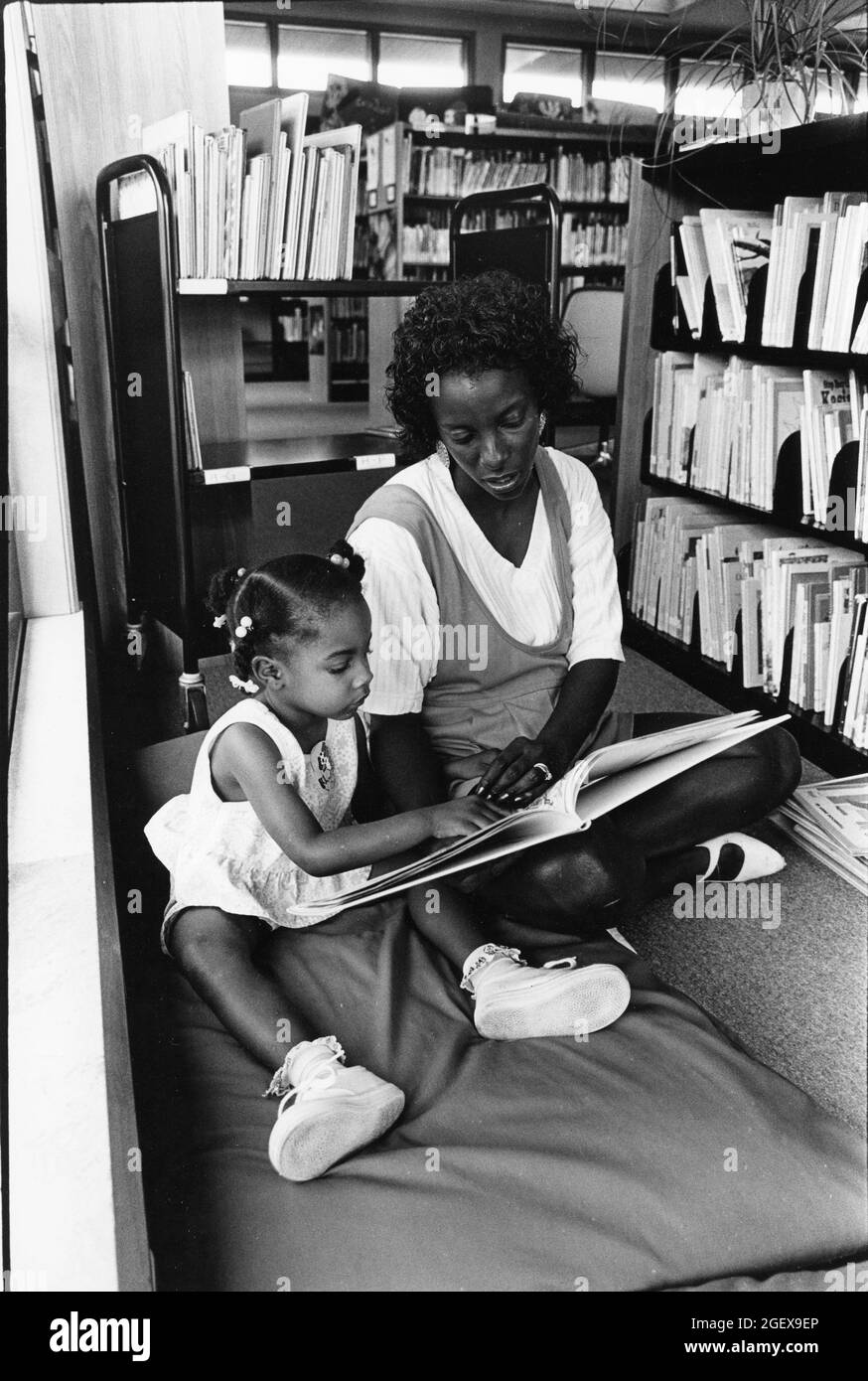 Austin Texas USA, circa 1992: Black mother and young daughter sit on floor and look at book together during a summertime reading program at a public library. ©Bob Daemmrich Stock Photo