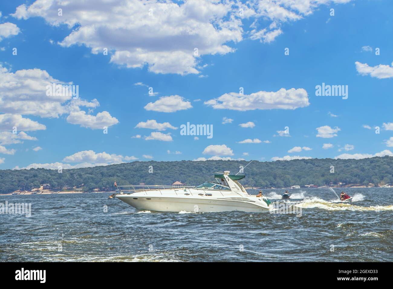 Motor yacht speeds across lake under pretty cloudy sky and riders of pwc jump the wake - shore with docks in distance Stock Photo