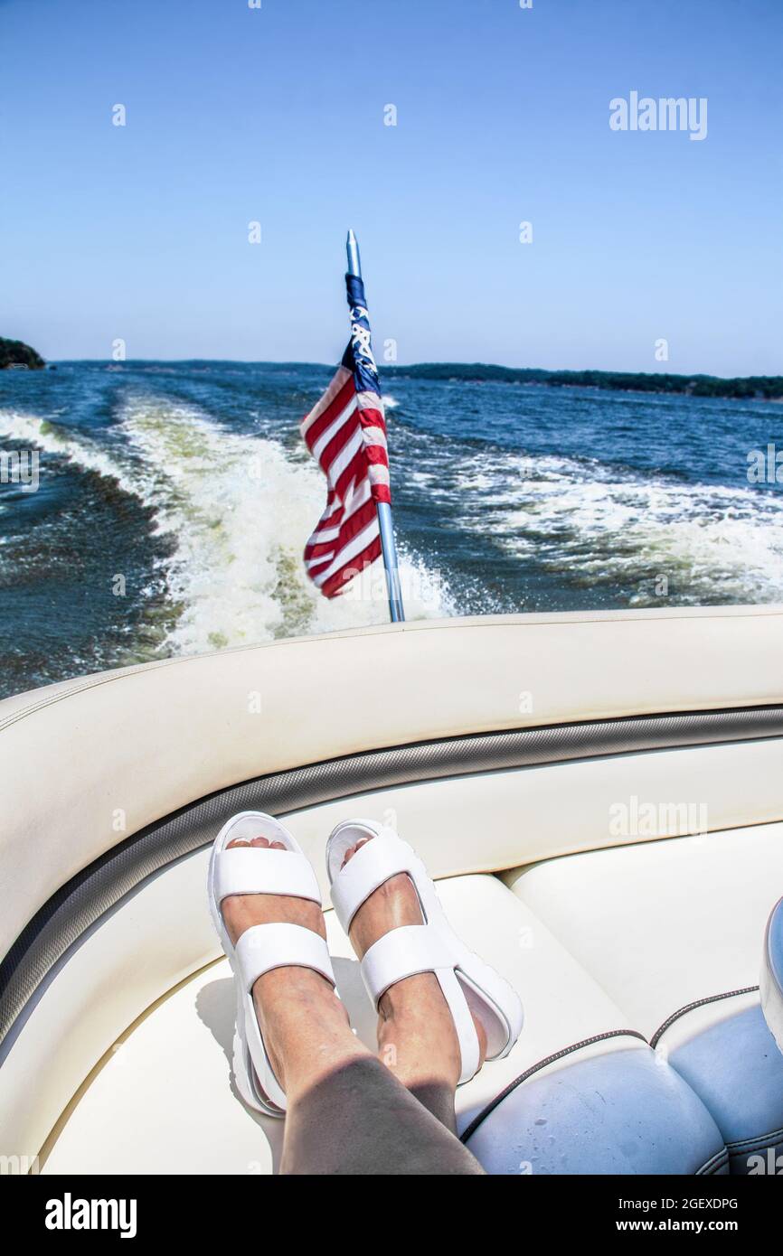Riding in the back of the speedboat across the lake - womens feet with white sandals propped on curved seat with flag and wake and lake whoosing by in Stock Photo