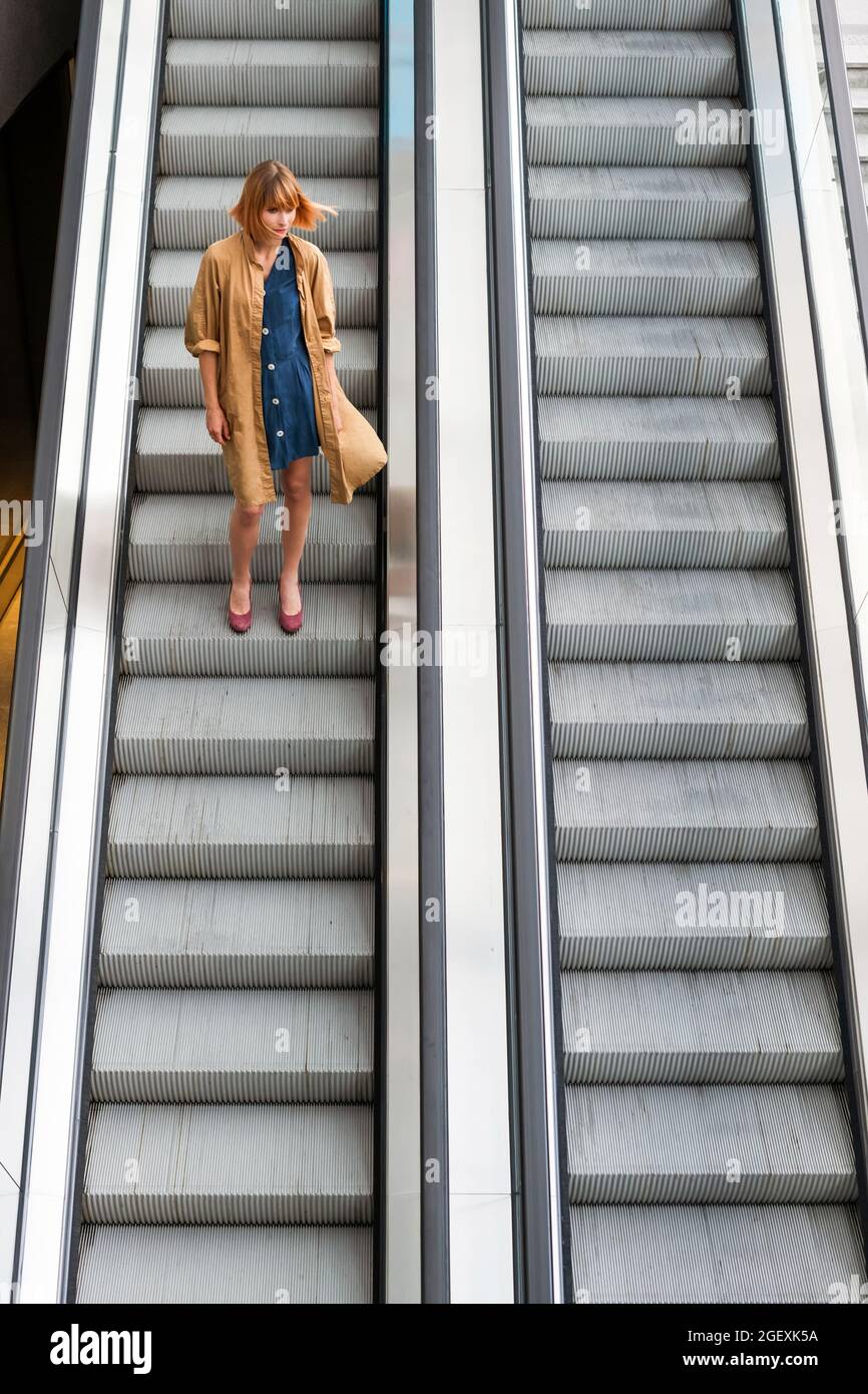 Trendy redhead woman riding alone on a steep long descending escalator in a city building or mall in a distance view with copyspace Stock Photo