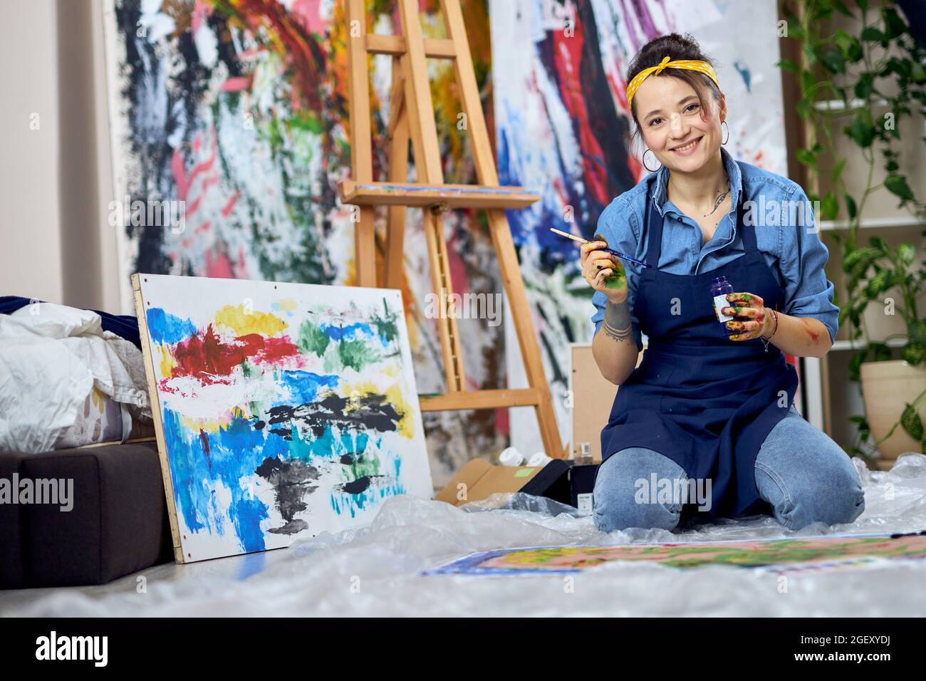 Joyful female painter in apron smiling at camera, holding paintbrush ...