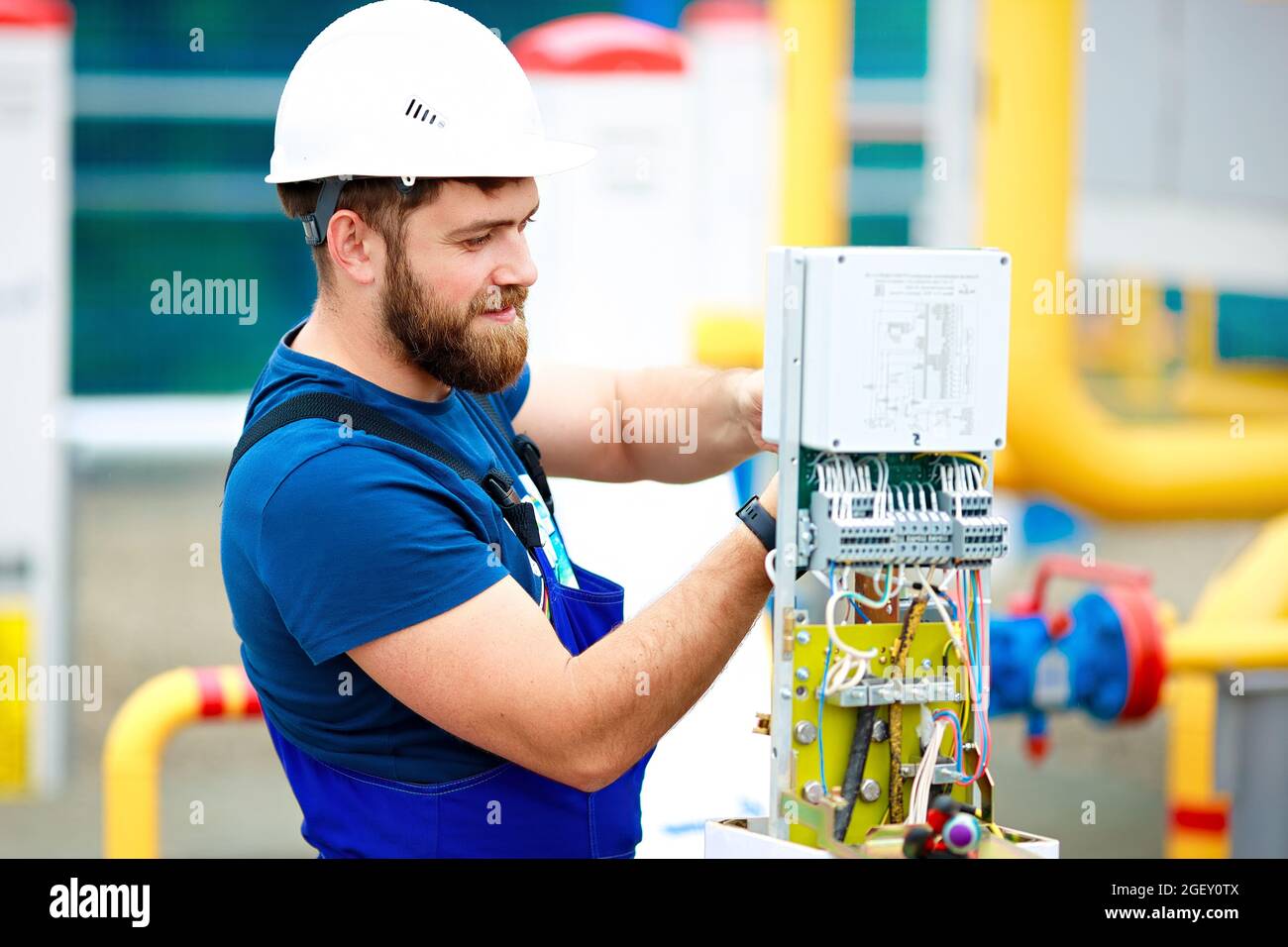 An engineer in a helmet and work clothes adjusts and inspects the equipment at the production facility. An authentic portrait of a Caucasian man with a beard at work. Stock Photo