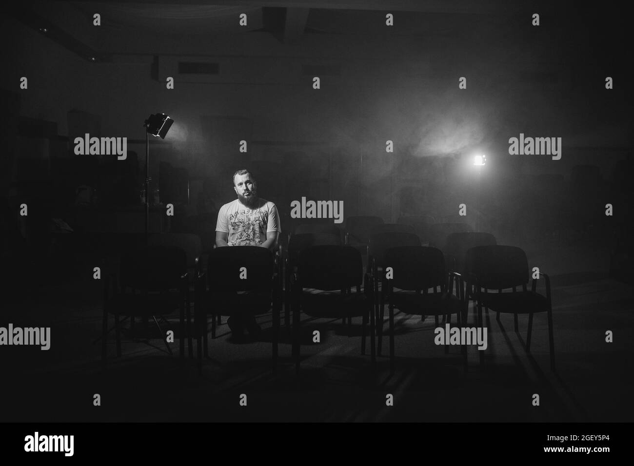 A grayscale shot of a Caucasian man sitting alone in an audience room with studio lights on him Stock Photo