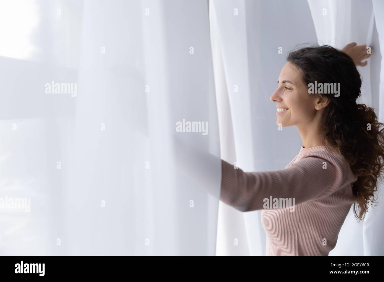 Young Latina woman open curtains looks out window feels happy Stock Photo