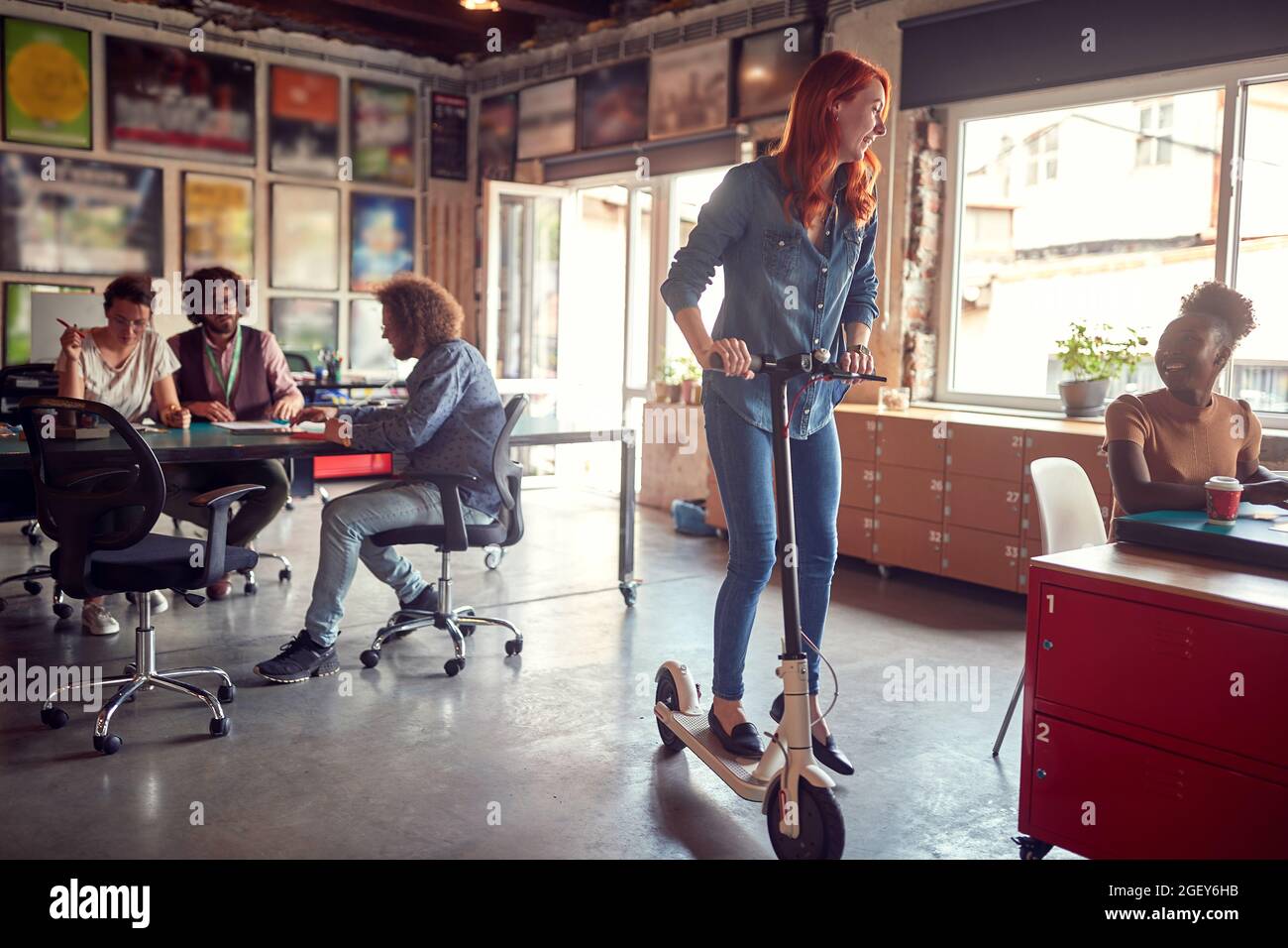 modern casual business approach concept. Young redhead on a scooter at work, taking a fun break, looking at her afro american female colleague, smilin Stock Photo
