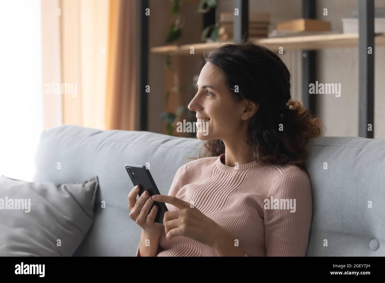 Smiling dreamy Latin woman sit on sofa holding modern cellphone Stock Photo