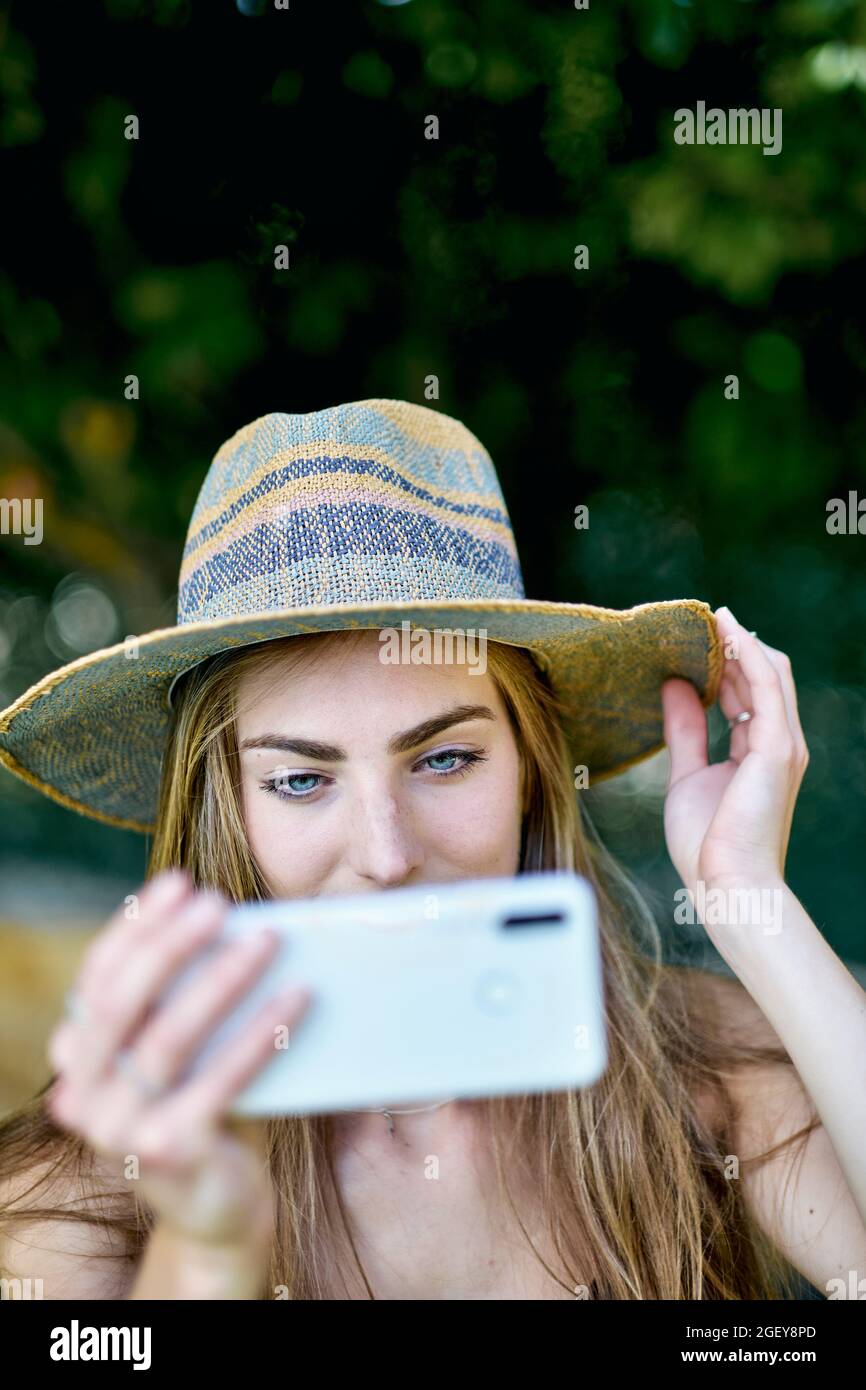 Natural beauty portrait of young woman in her 20s with long hair and blue eyes outdoor in a garden wearing a hat and shotting a selfie with her mobile Stock Photo