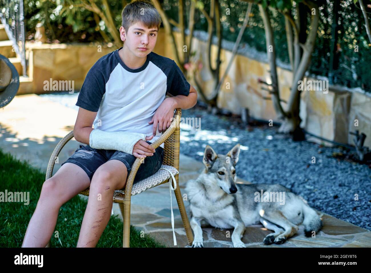 Portrait of young caucasian boy with a broken and cast arm sitting in a chair outdoor in a garden with a dog. Lifestyle concept. Stock Photo