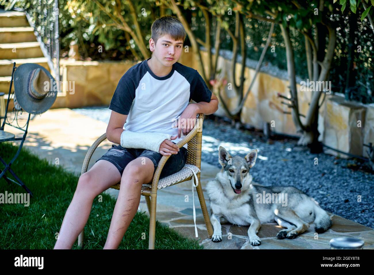 Portrait of young caucasian boy with a broken and cast arm sitting in a chair outdoor in a garden with a dog. Lifestyle concept. Stock Photo