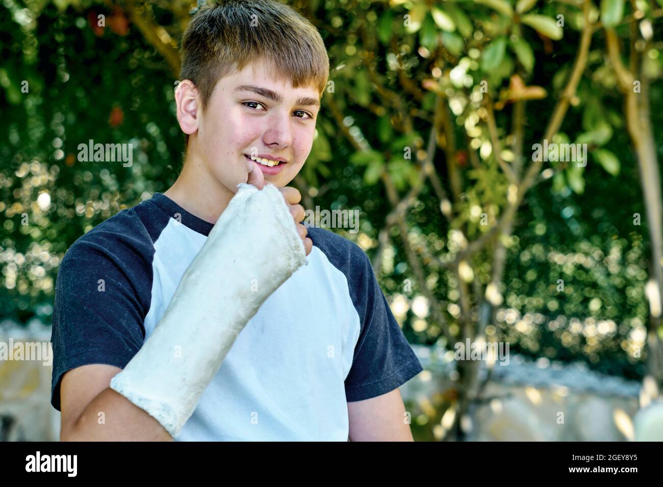 Portrait of young caucasian boy with a broken and cast arm sitting in a chair outdoor in a garden. Lifestyle concept. Stock Photo