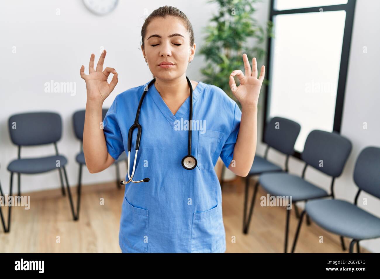 Young brunette doctor woman at waiting room relax and smiling with eyes closed doing meditation gesture with fingers. yoga concept. Stock Photo