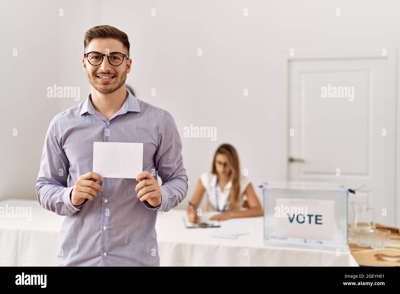 Young voter man holding blank vote standing by electoral table at vote center. Stock Photo