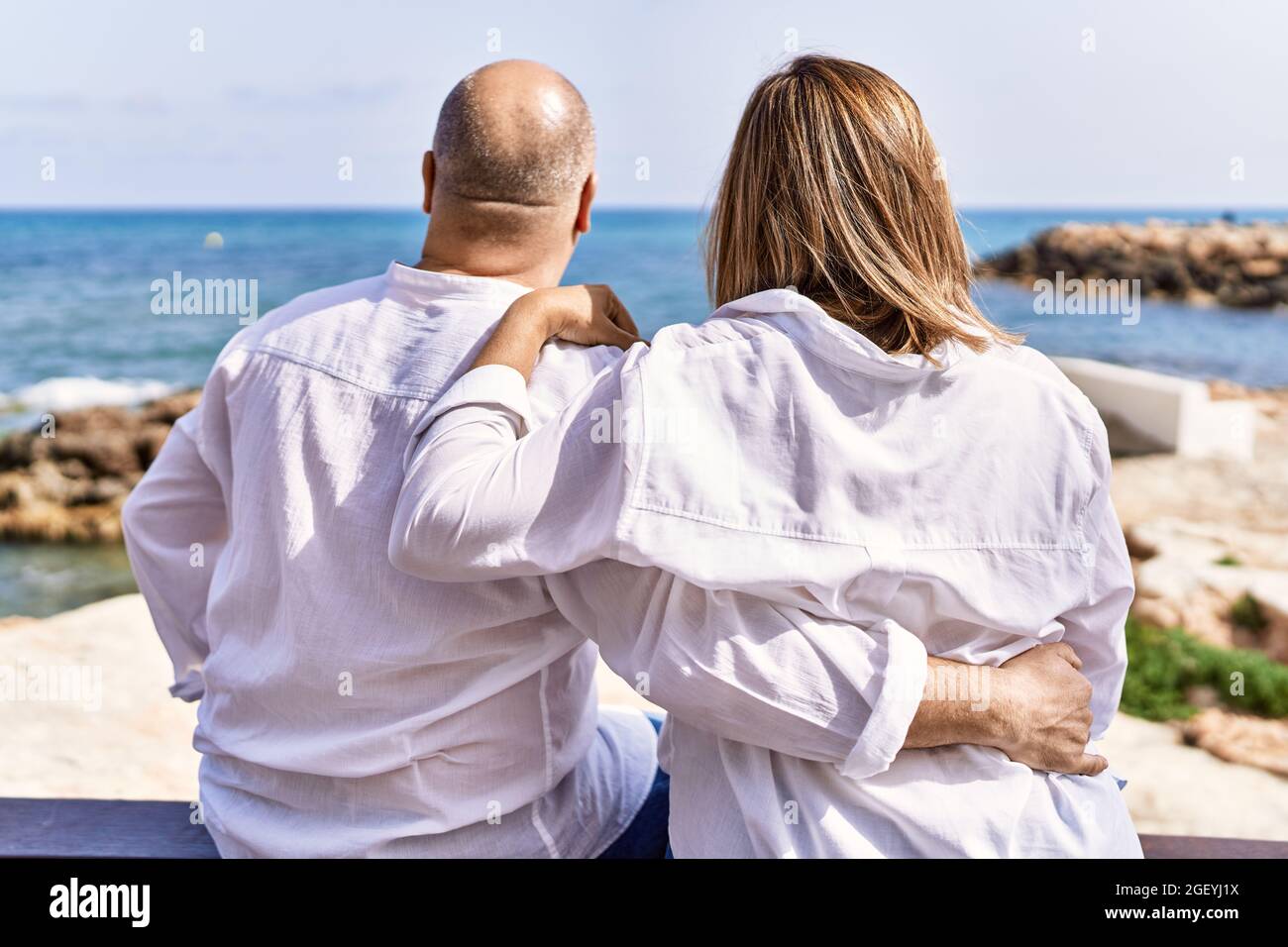 Middle age hispanic couple of husband and wife together sitting by the beach on a sunny day