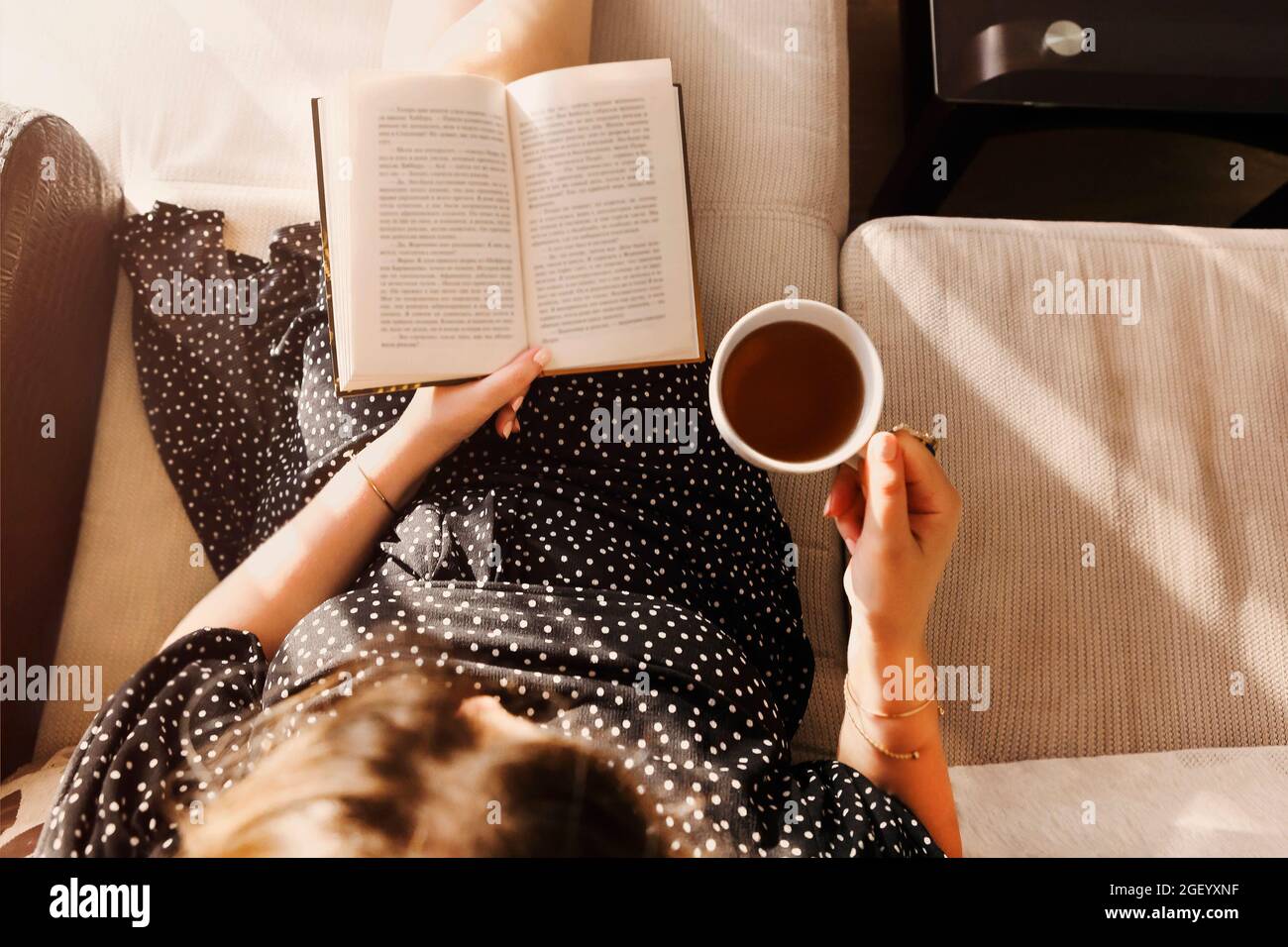 Photo from above of young woman in casual dress sitting with book and cup of tea on cozy couch at home, relaxed happy lady spending leisure time in si Stock Photo