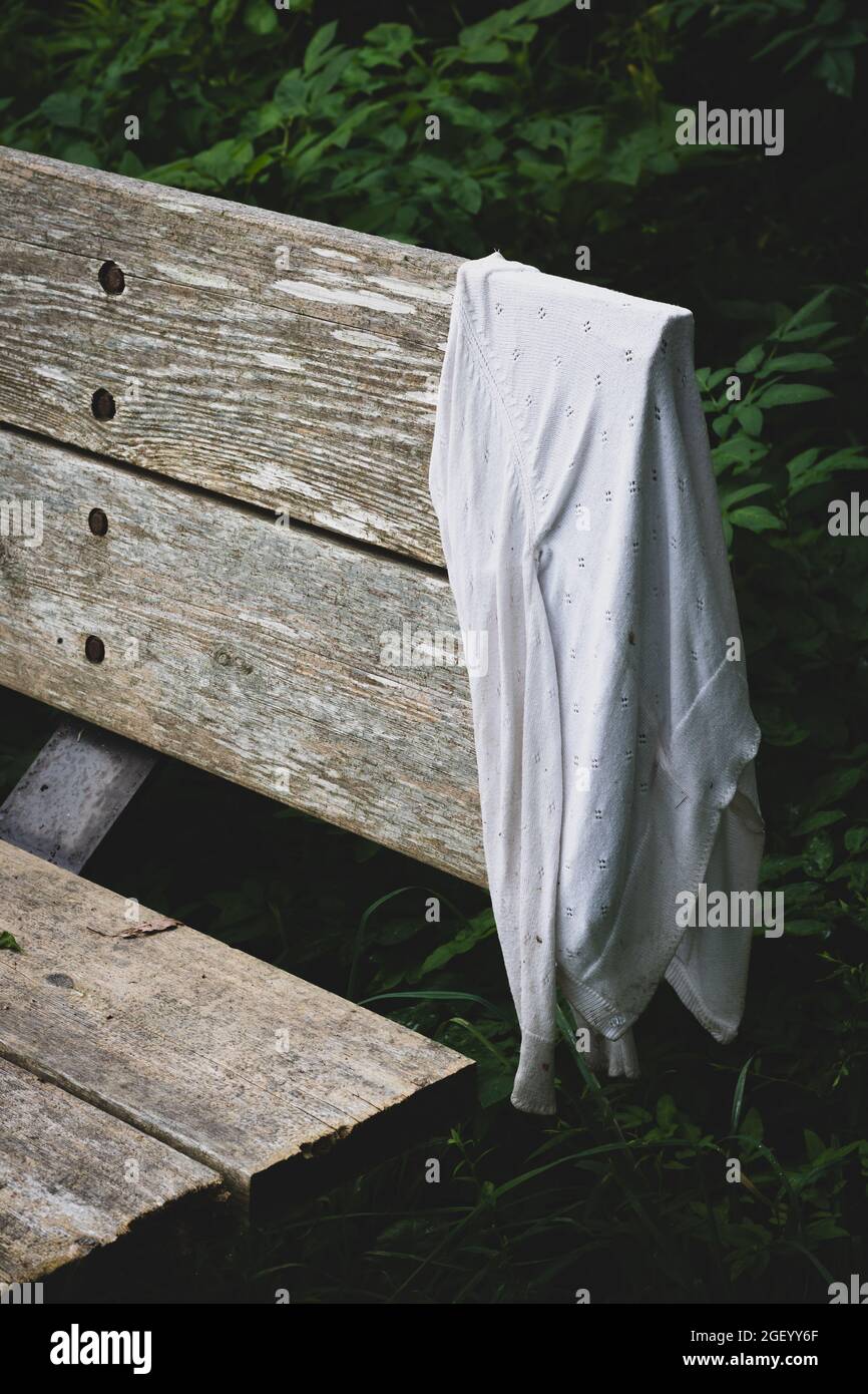 Selective focus of lost women's white cardigan sweater hung on wooden bench on hiking trail. Grief and loss concept. Stock Photo