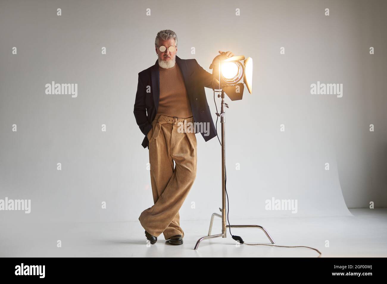 Full length shot of stylish grey haired middle aged man in glasses looking at camera, standing next to studio spotlight while posing over white Stock Photo