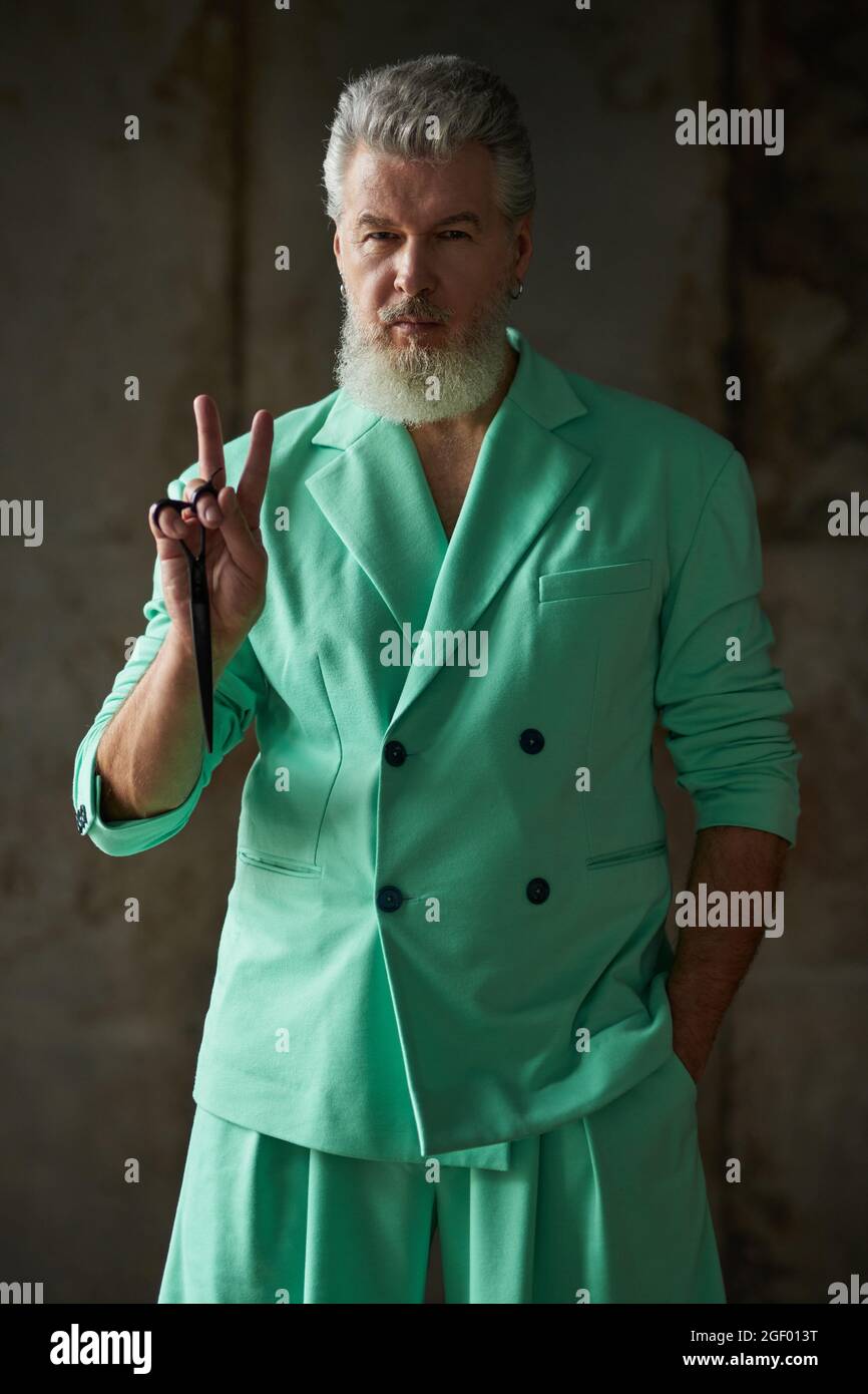 Cool stylish gray haired mature man with beard wearing colorful outfit looking at camera, showing peace sign while holding sharp barber scissors Stock Photo