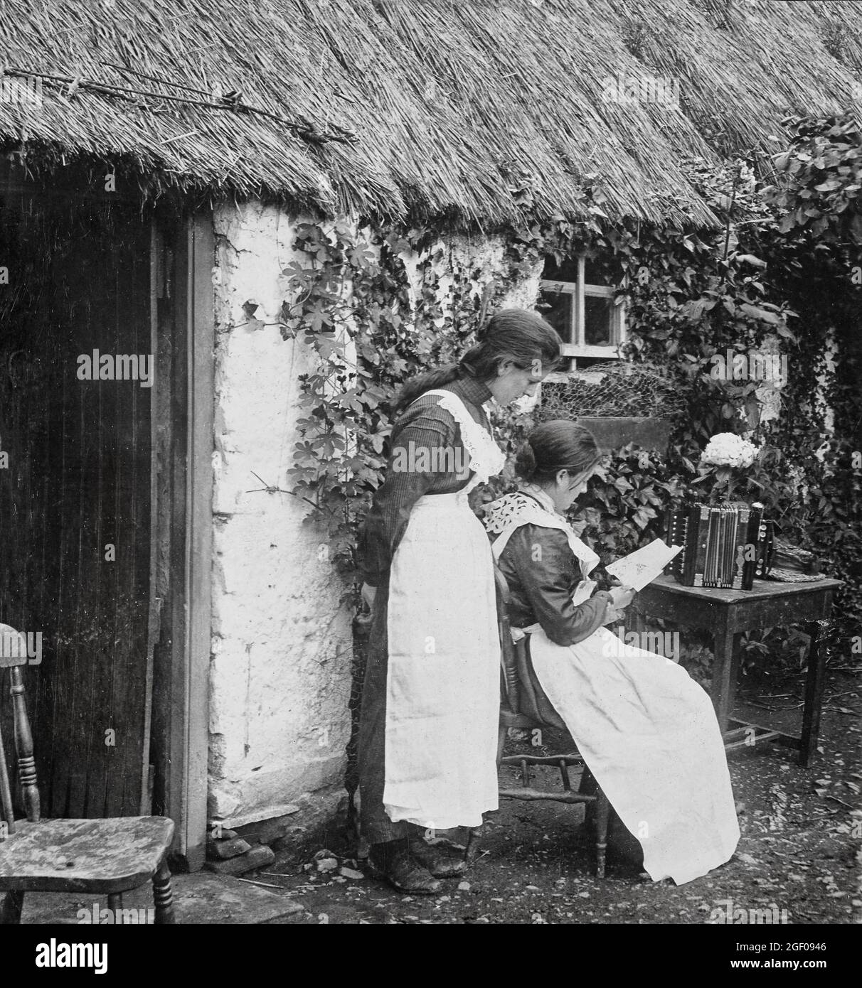 Two young ladies interrupt their accordion playing to read a newly arrived letter outside their cottage in early 20th century County Monaghan, Ireland Stock Photo