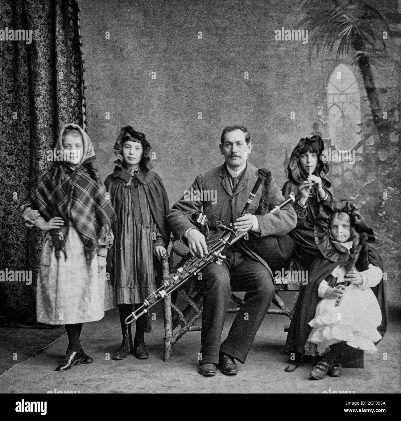 An early 20th century portrait of a musical family in County Cork, Ireland. The daughters on the left are ready to dance another is playing a penny whistle. The young girl on the right is playing simplified Uilleann pipes while father plays a full set. Stock Photo