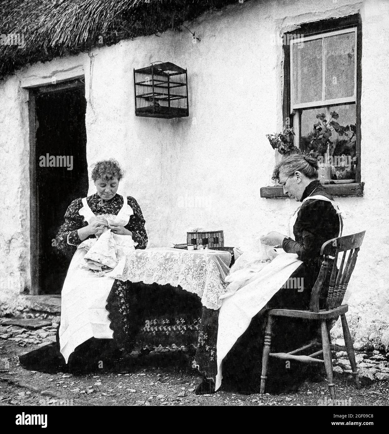 An early 20th century scene of two women making Carrickmacross Lace outside their thatched cottage in County Monaghan. It was introduced into Ireland in about 1820 by one Mrs Grey Porter of Donaghmoyne, who taught it to local women so that they could earn some extra money. Porter had been inspired by some examples of appliqué lace she had seen while on her honeymoon in Italy. Stock Photo