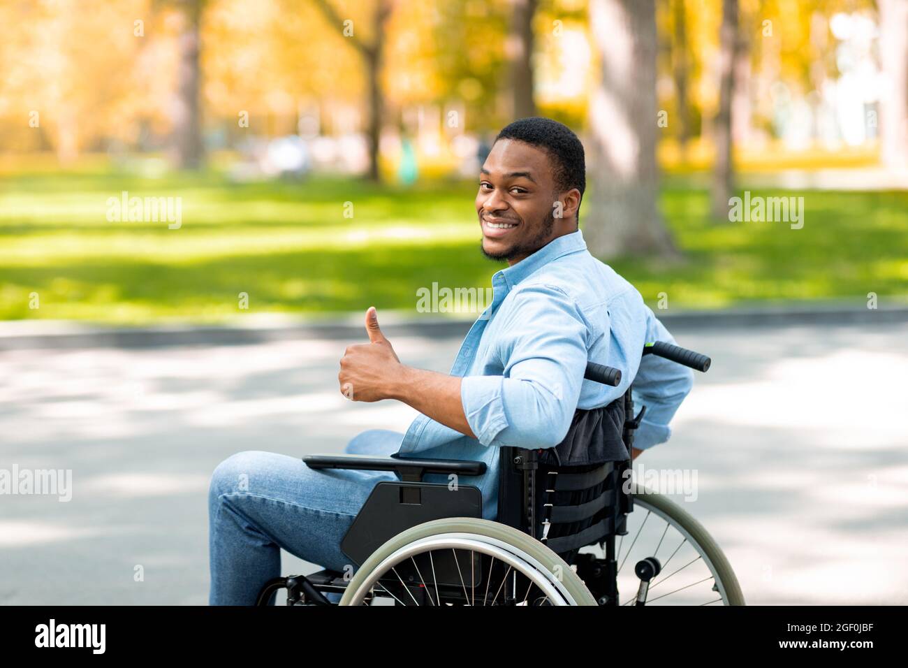 Happy disabled black man in wheelchair spending time at city park in autumn, showing thumb up gesture and smiling Stock Photo