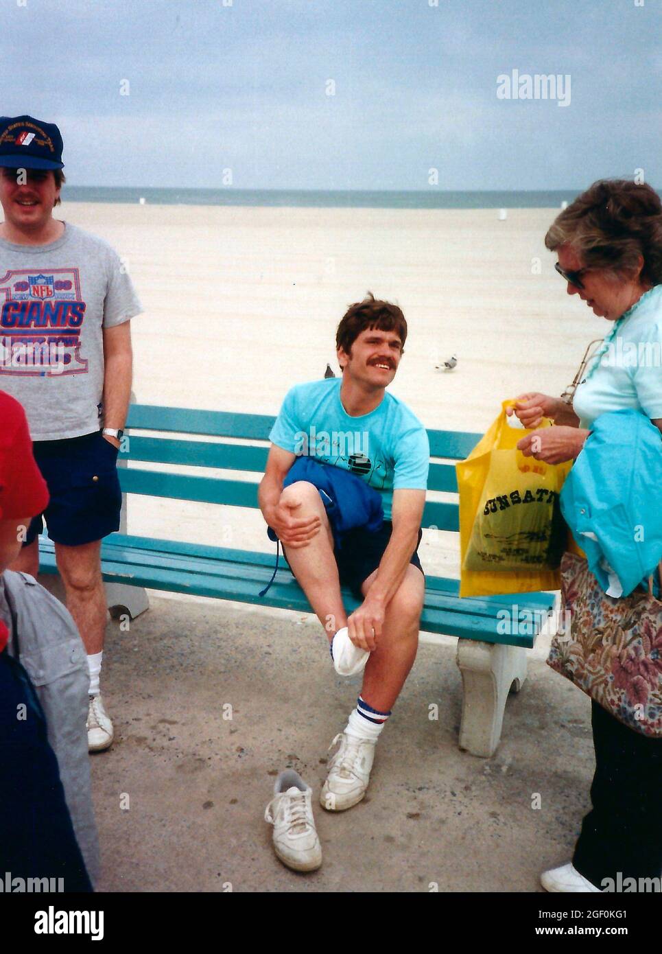 Man sitting on a bench and taking off his shoes while others stand around.  On the boardwalk in Ocean City, Maryland, circa 1988 Stock Photo