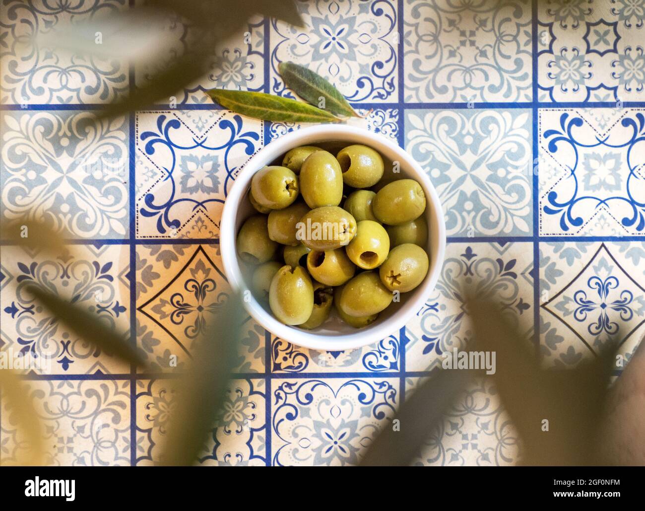 Small white bowl with a green olives snack on a table with traditional Mediterranean blue pattern tiles under an olive tree Stock Photo
