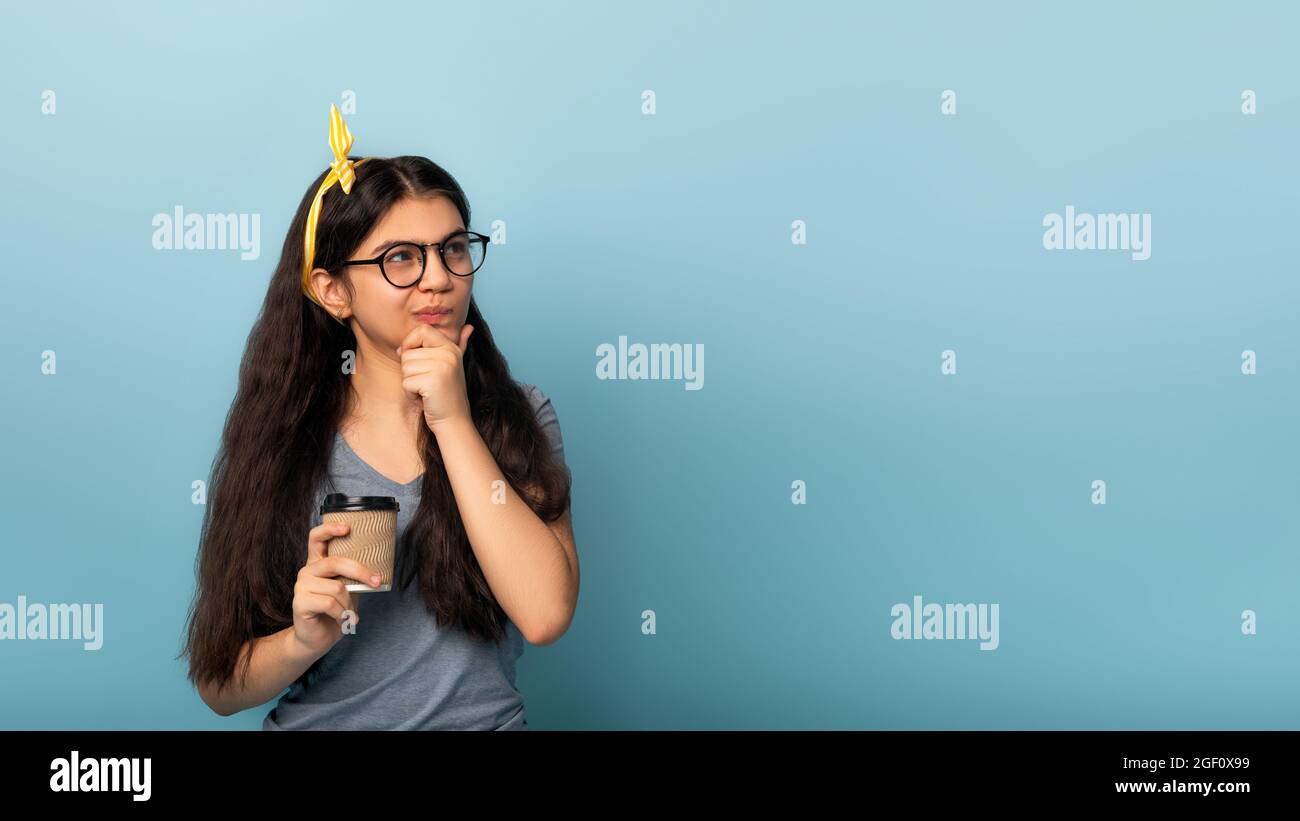 Pensive Indian teen girl holding cup of takeaway coffee, pondering on proposal, deep in thought on blue background Stock Photo